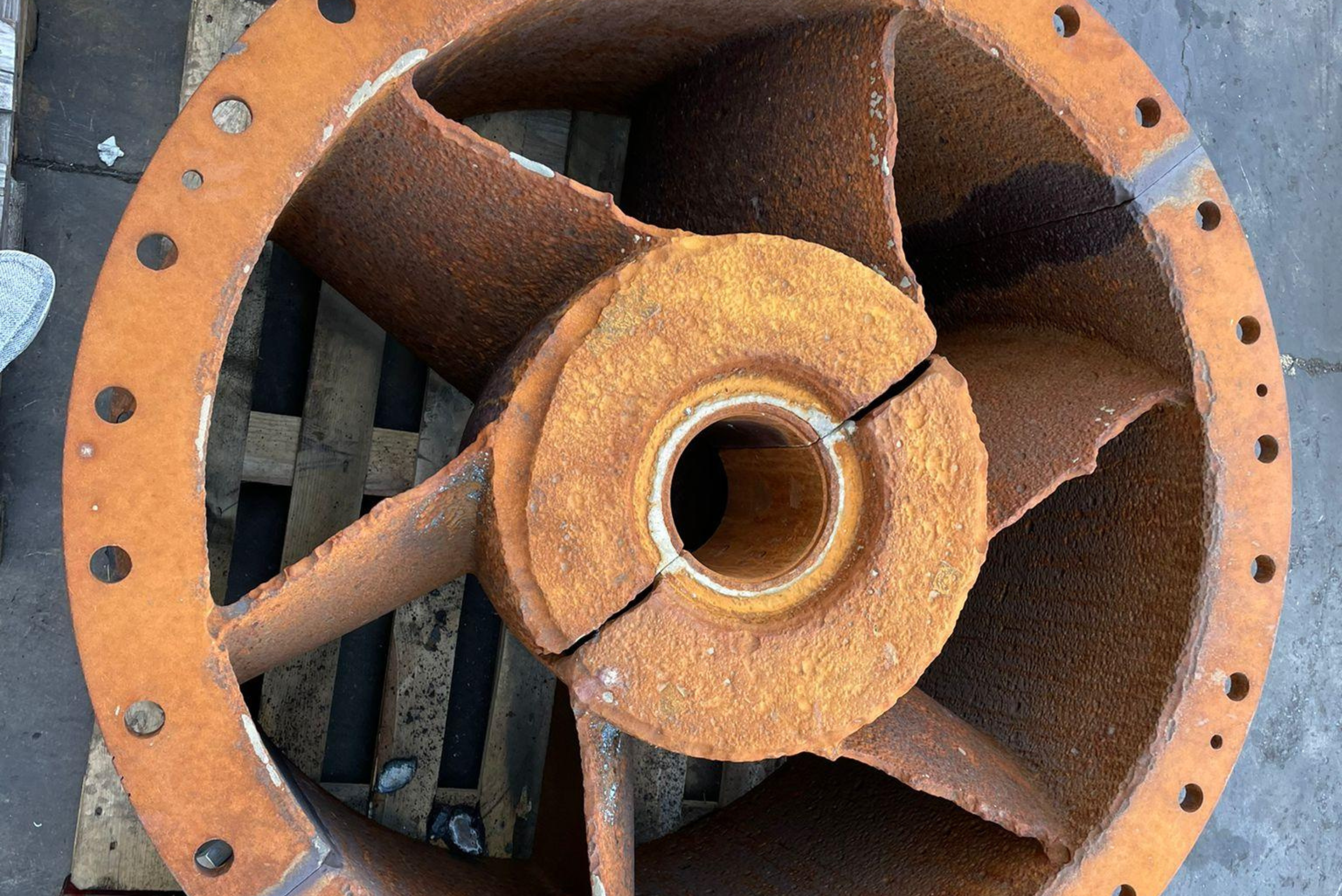 Top-down view of a circular, heavily corroded metallic impeller or pump component with damaged blades and a visible crack through the central hub, resting on a wooden pallet.