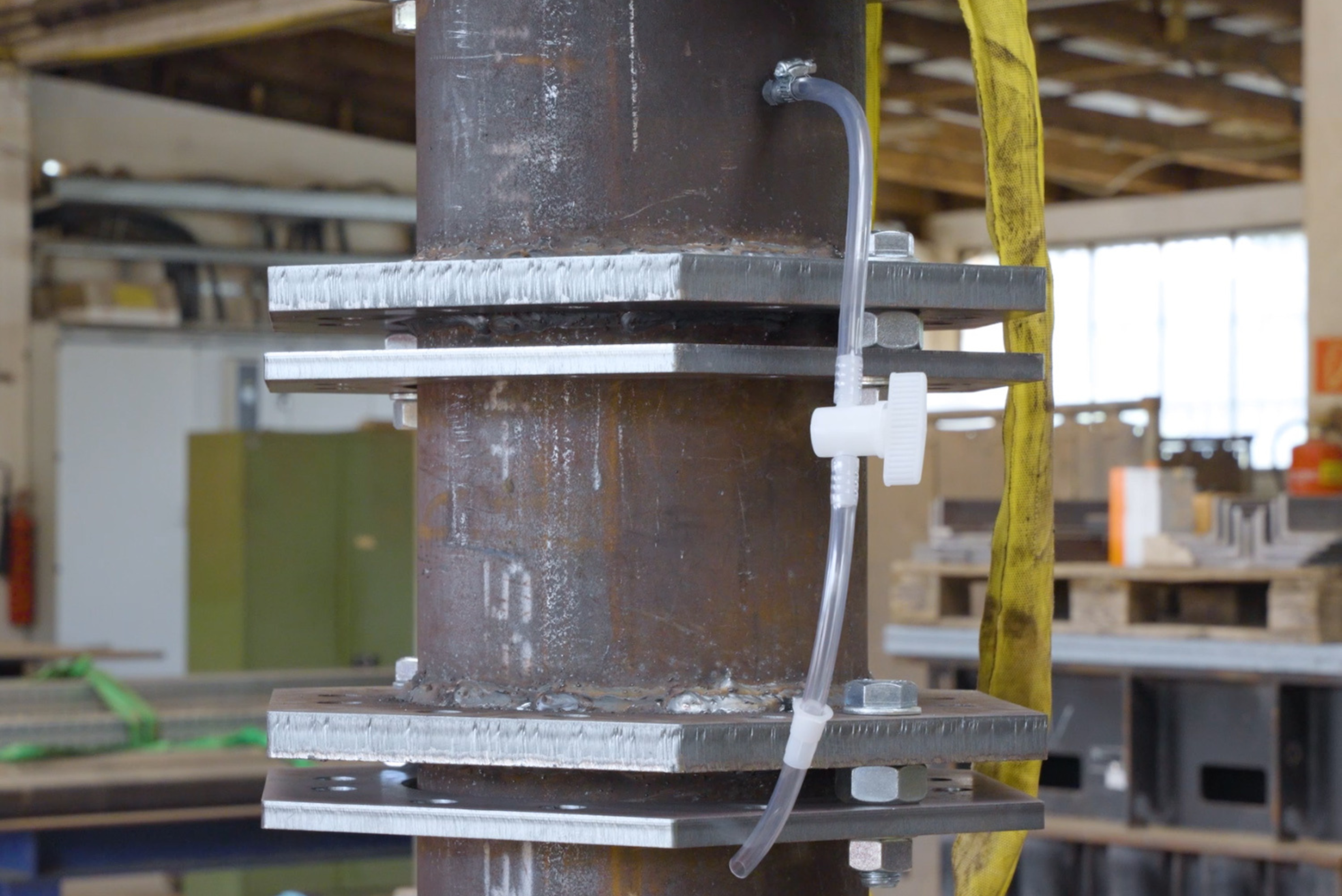 Vertical steel pipe assembly with bolted flange connections, featuring a clear flexible hose attached with a small valve for venting or fluid transfer.