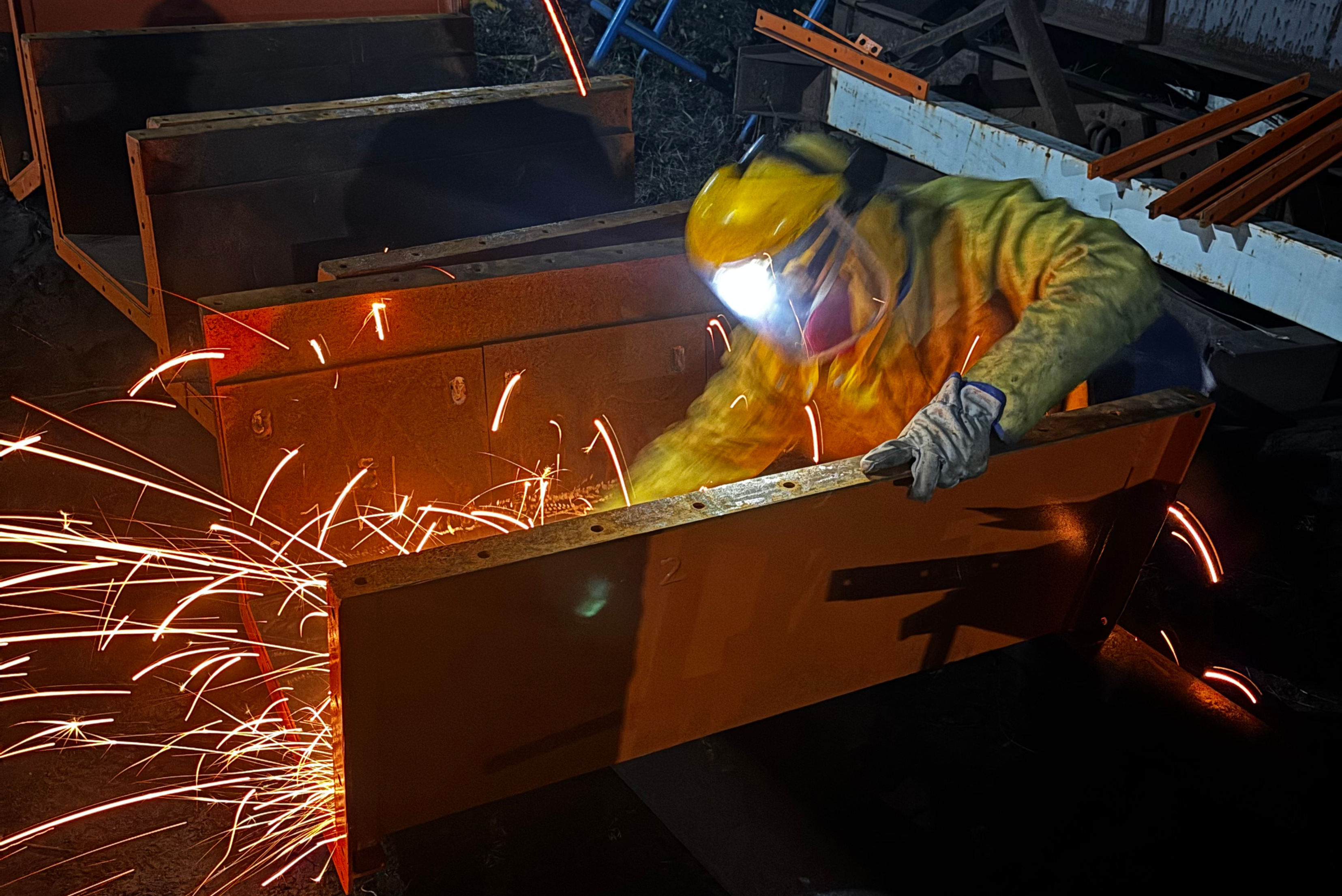 Industrial worker wearing protective gear, a helmet with a face shield, and a respirator, grinding heavy metal components with a power tool while flying sparks illuminate the dark workshop surroundings.