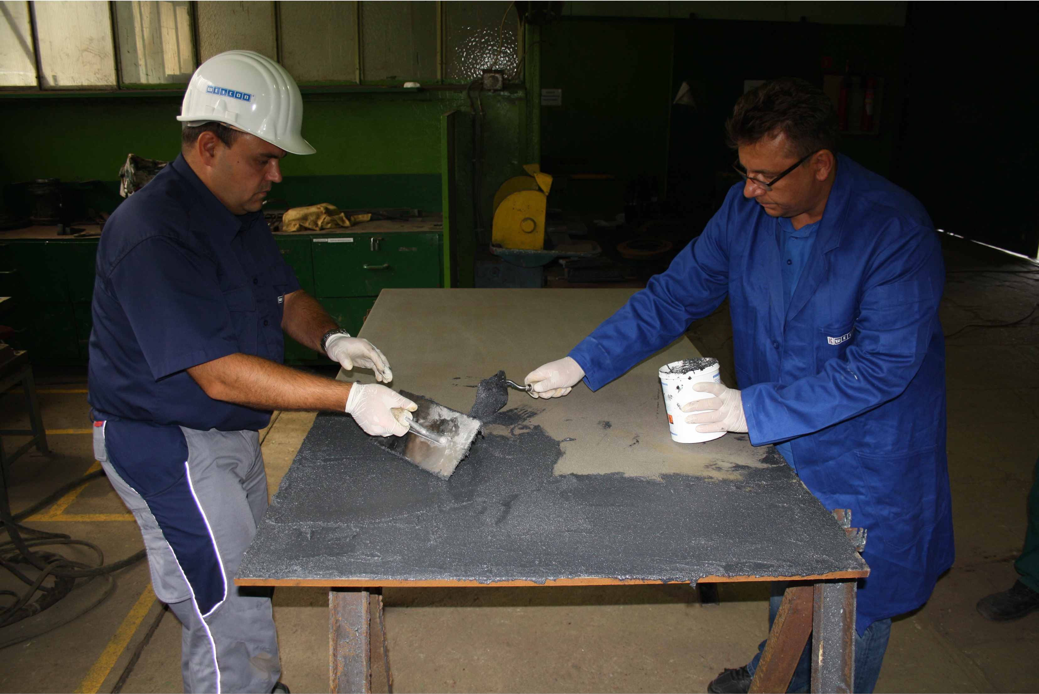 Two industrial technicians wearing protective gloves applying a thick grey coating compound onto a large flat steel plate using trowels in a workshop setting