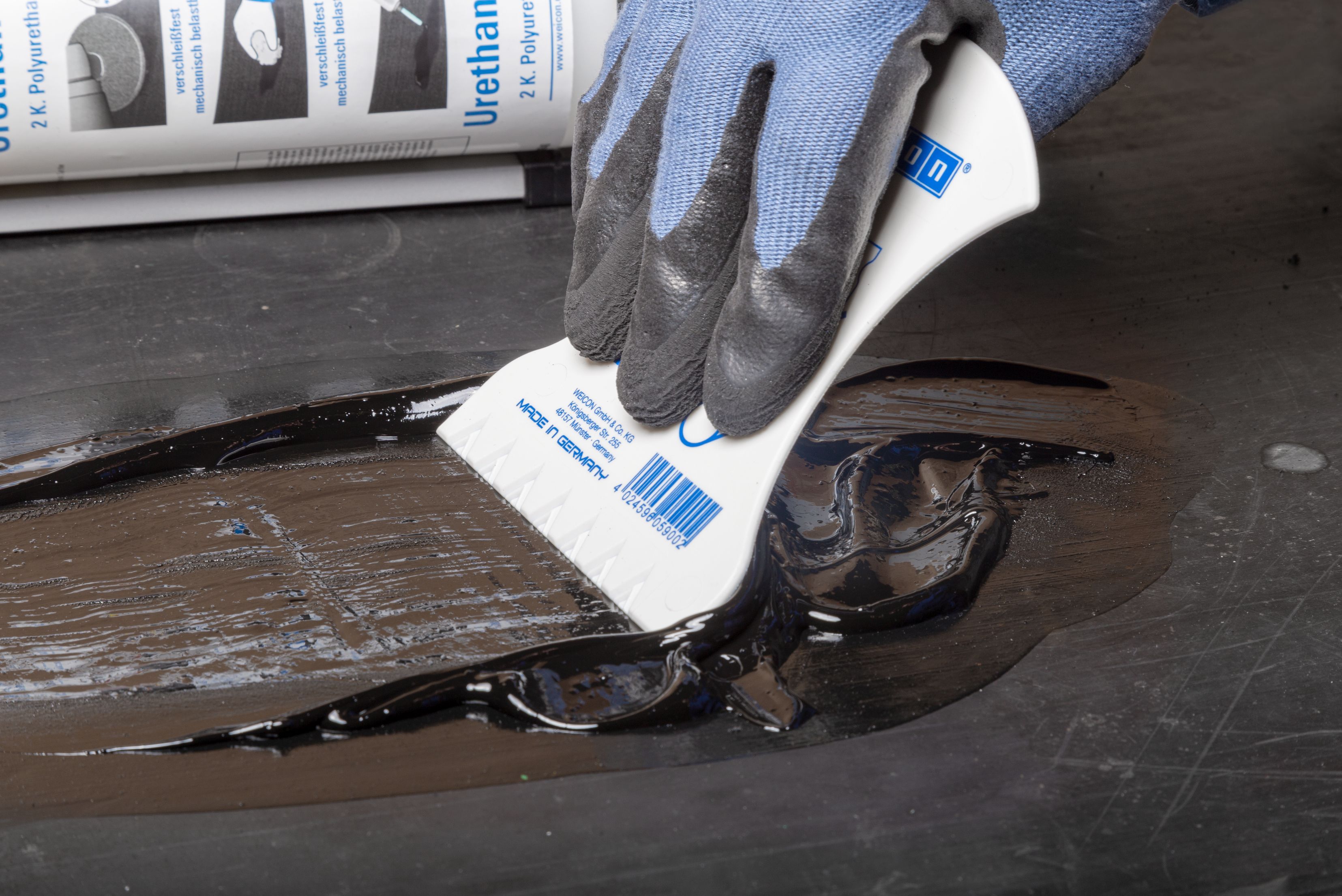 Extreme close-up of a worker in protective gloves using a white plastic contoured spatula to smooth and level a liquid black urethane repair resin over a damaged section of a dark conveyor belt, with the compound cartridge visible in the background.