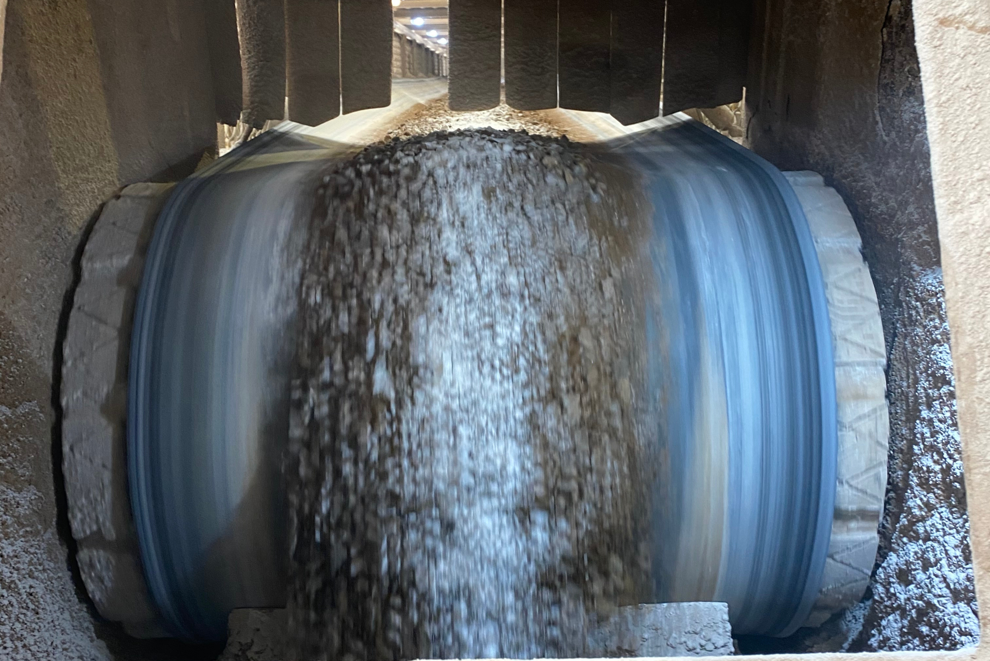 A close-up view of a rotating industrial conveyor belt pulley transporting bulk material, such as crushed stone or aggregate, through a enclosed processing facility.