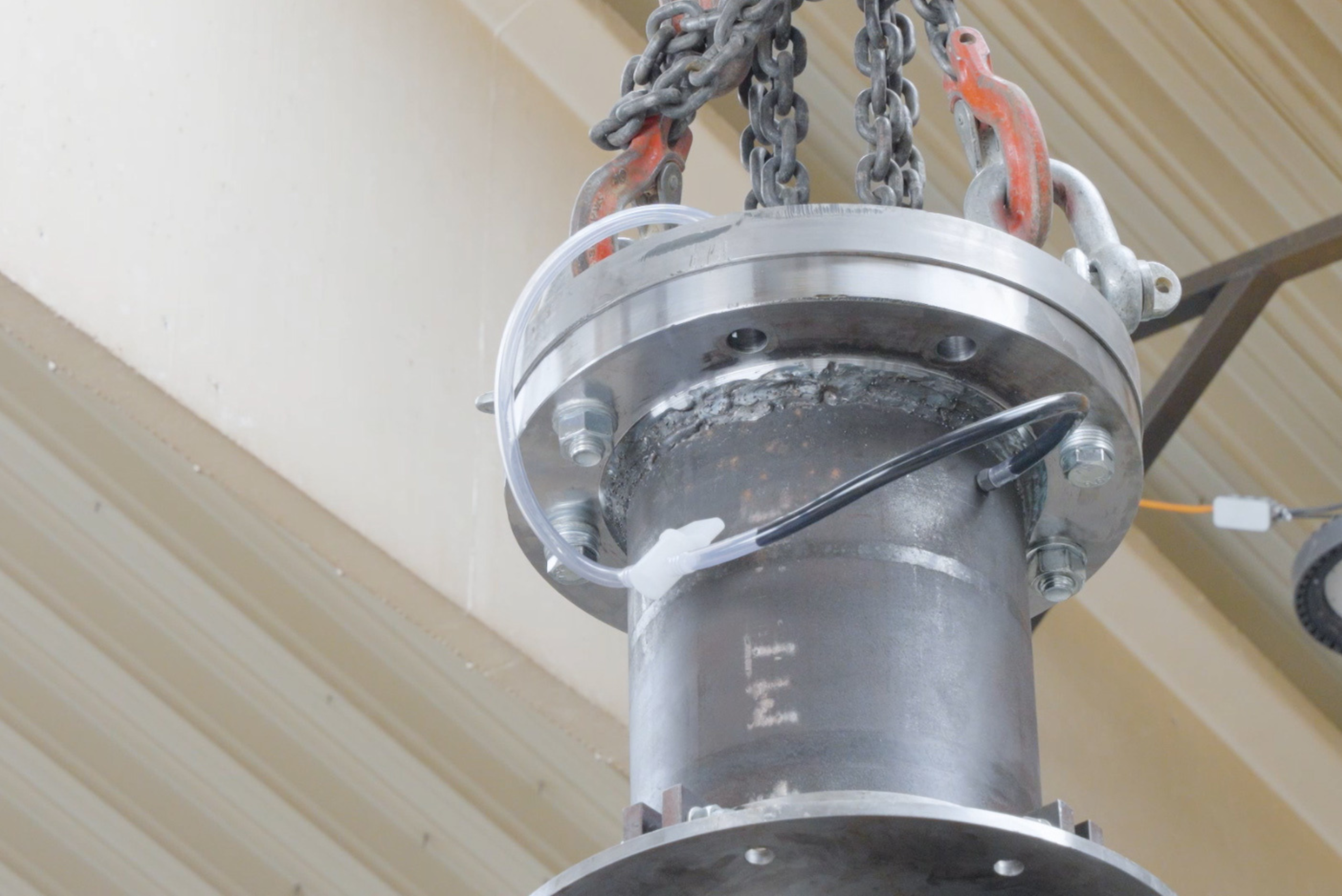 Vertical steel pipe segment with heavy flange connections, suspended by chain slings and shackles, equipped with connected fluid transfer and venting lines.