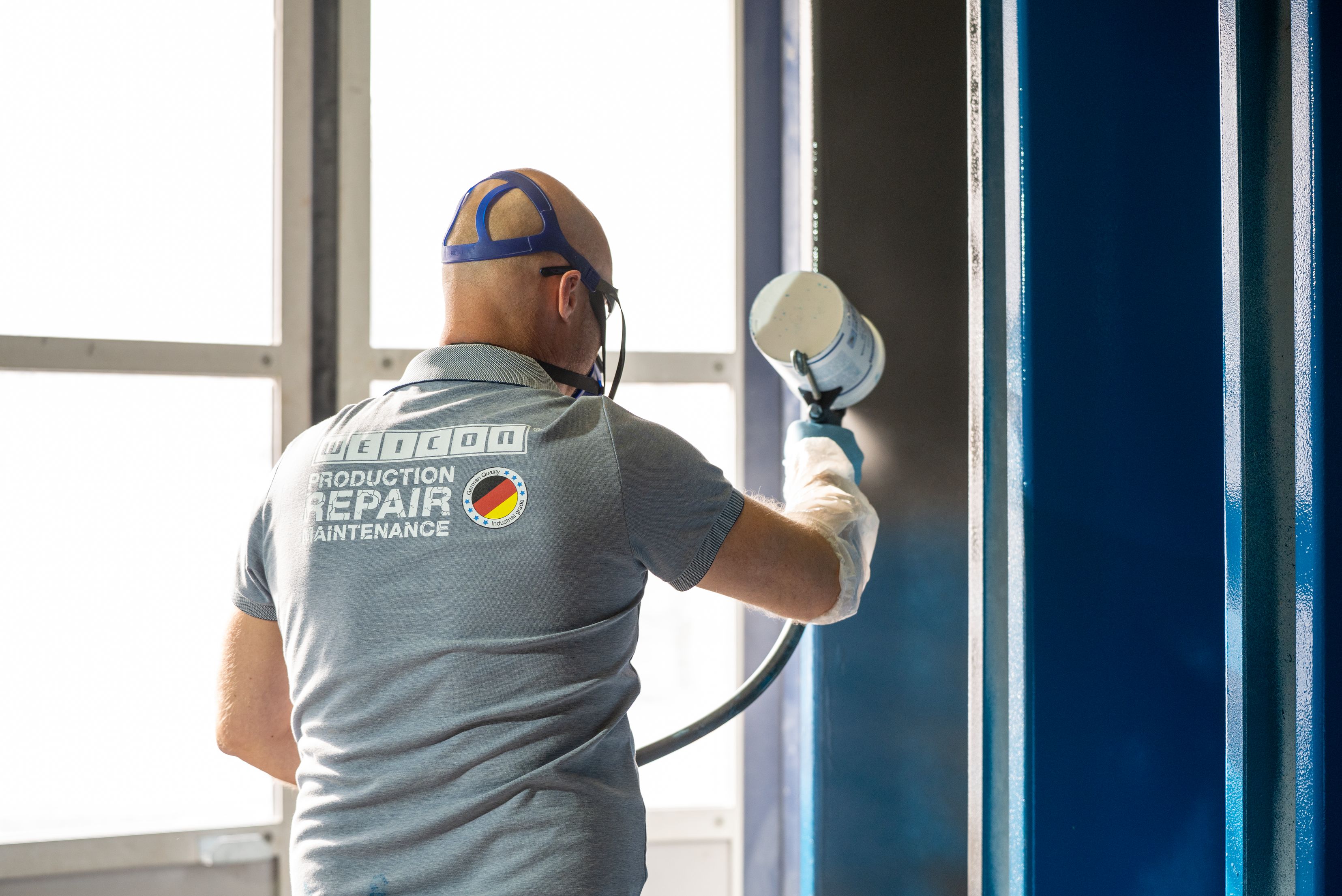 Rear view of a worker wearing protective glasses, a respirator mask, and protective arm sleeves using a spray gun to apply a protective coating to a vertical metallic structure in a workshop area
