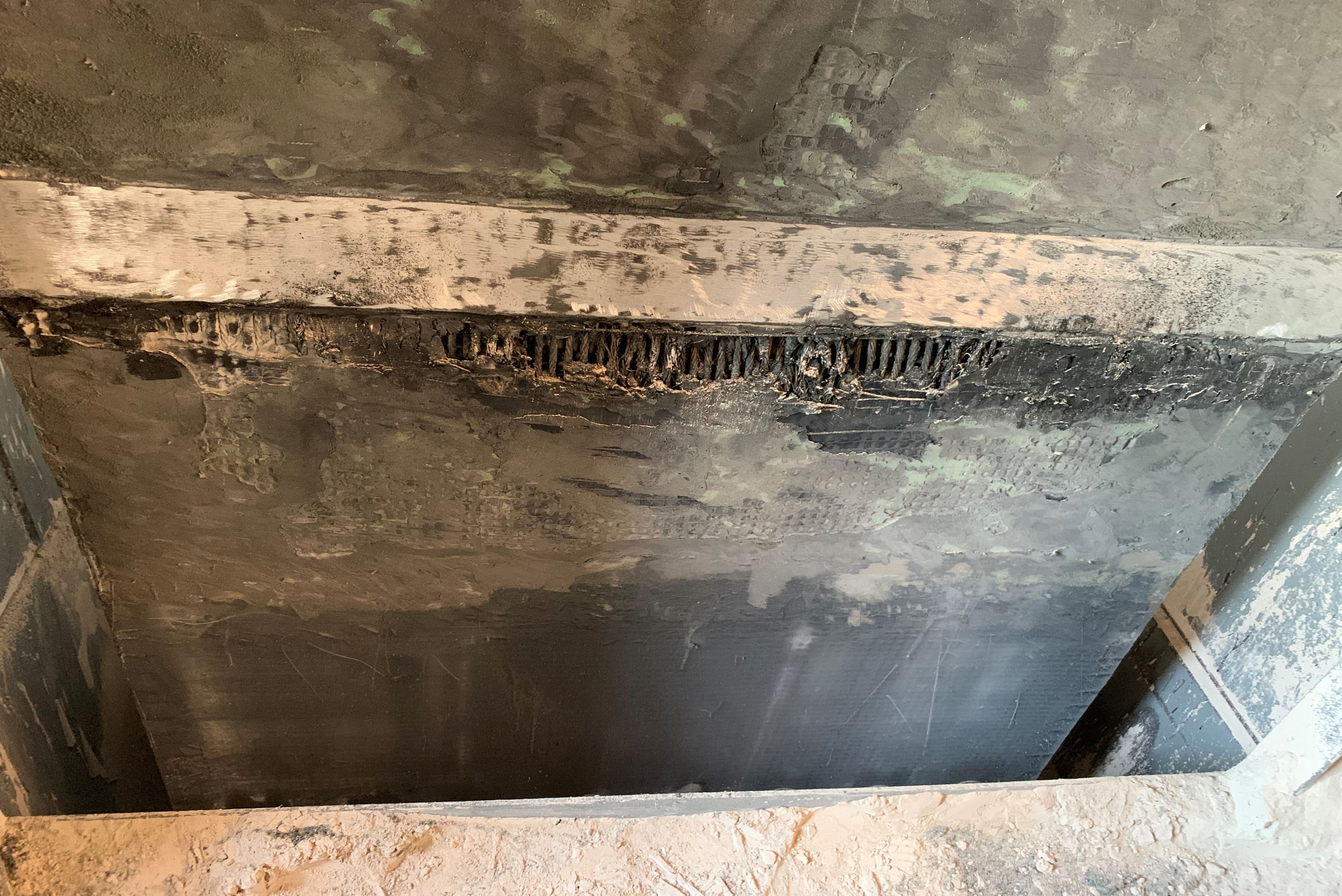 Underneath or interior view of a concrete slab or beam showing severe surface degradation and exposed, corroded rebar mesh along the joint edge.