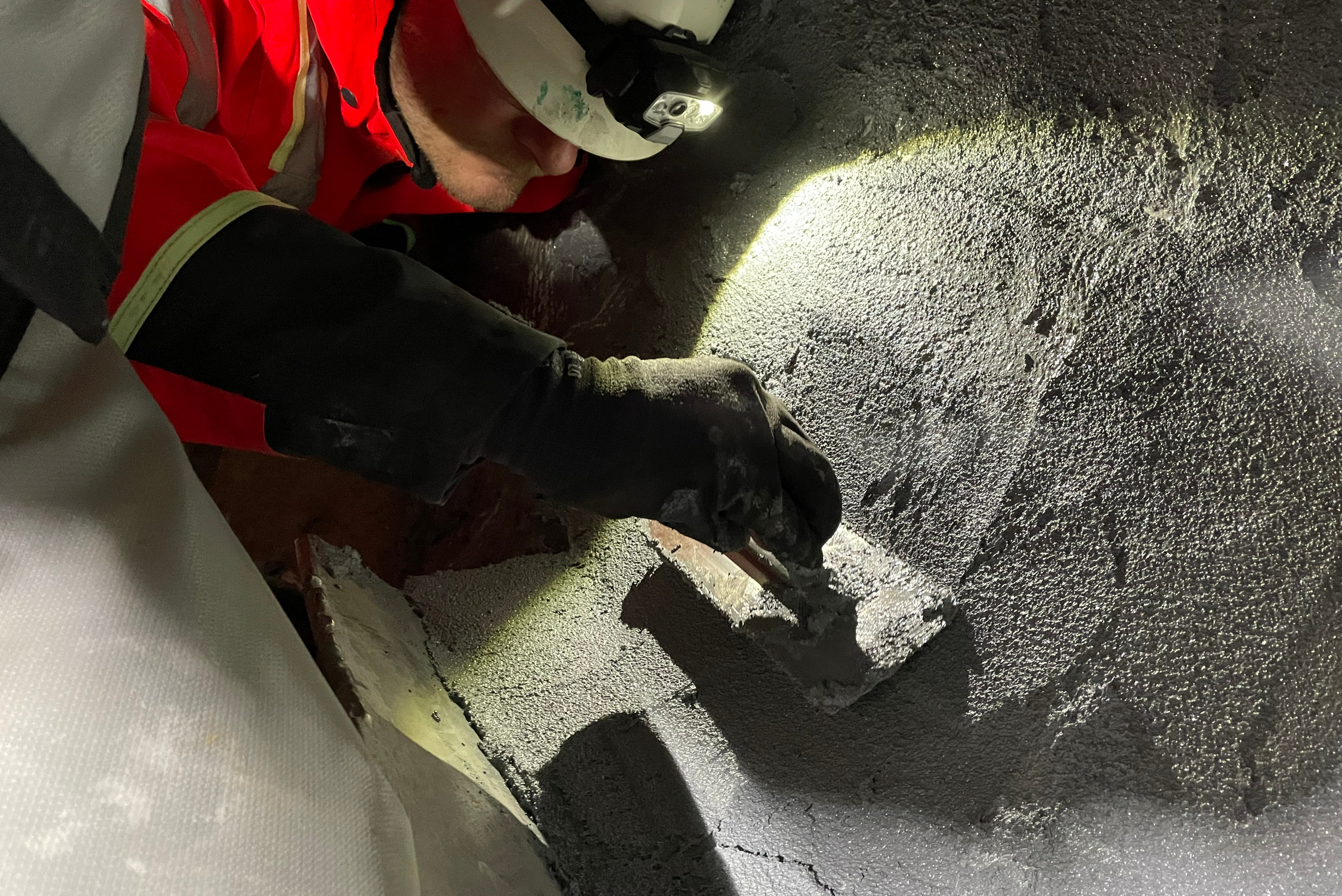 Close-up view of a technician wearing a hardhat with a headlamp, using a trowel to apply a thick grey protective coating to the rough interior surface of an industrial vessel or chamber.