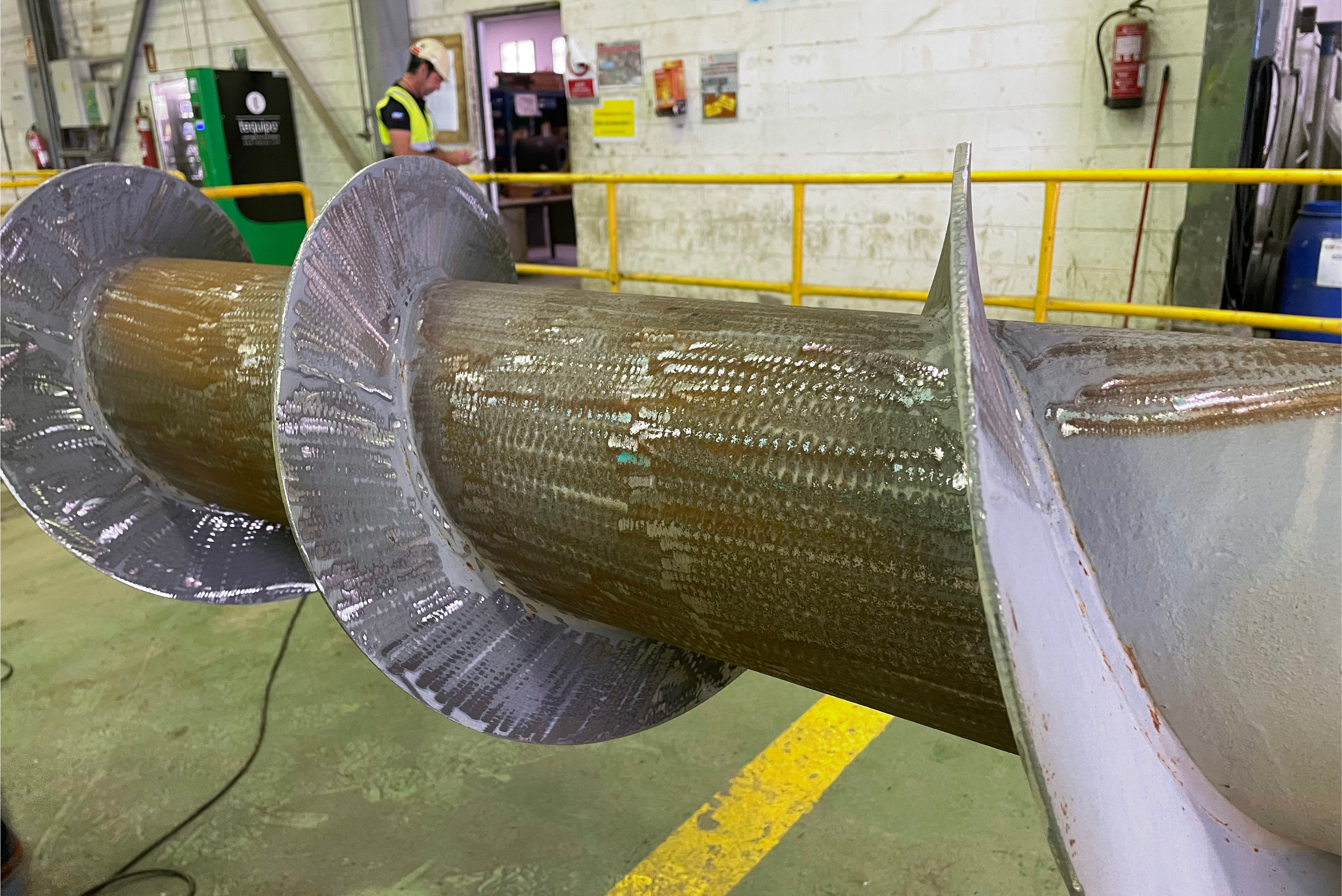 Close-up view of a large screw conveyor with a visible protective coating applied to the flights and central shaft, positioned inside a workshop facility with a worker standing in the background.