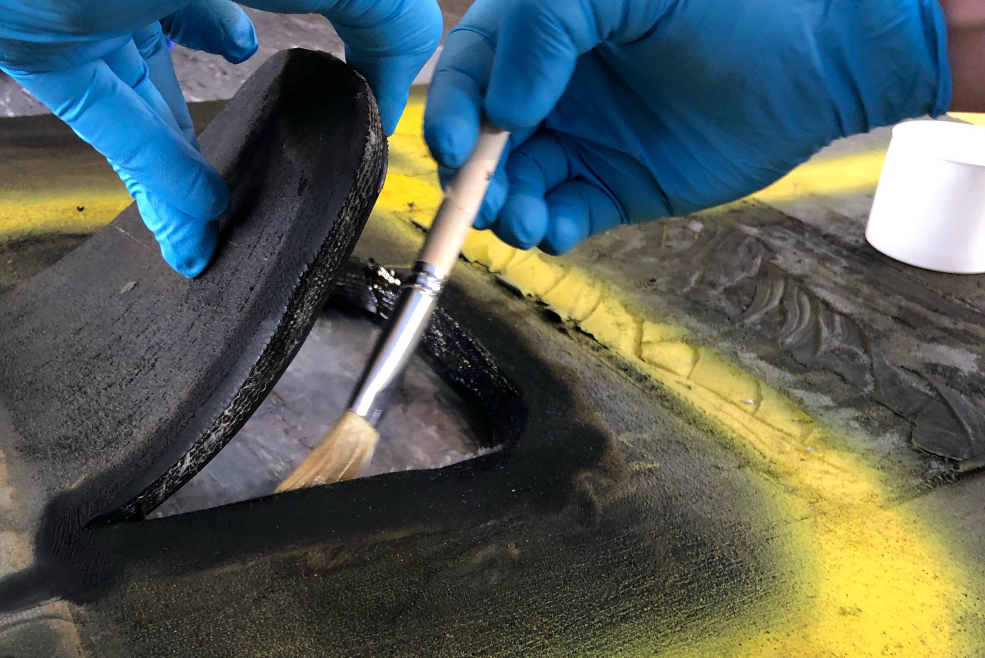 Close-up of a person wearing blue gloves using a paintbrush to apply black adhesive or sealant around a fitted component on a dark surface.