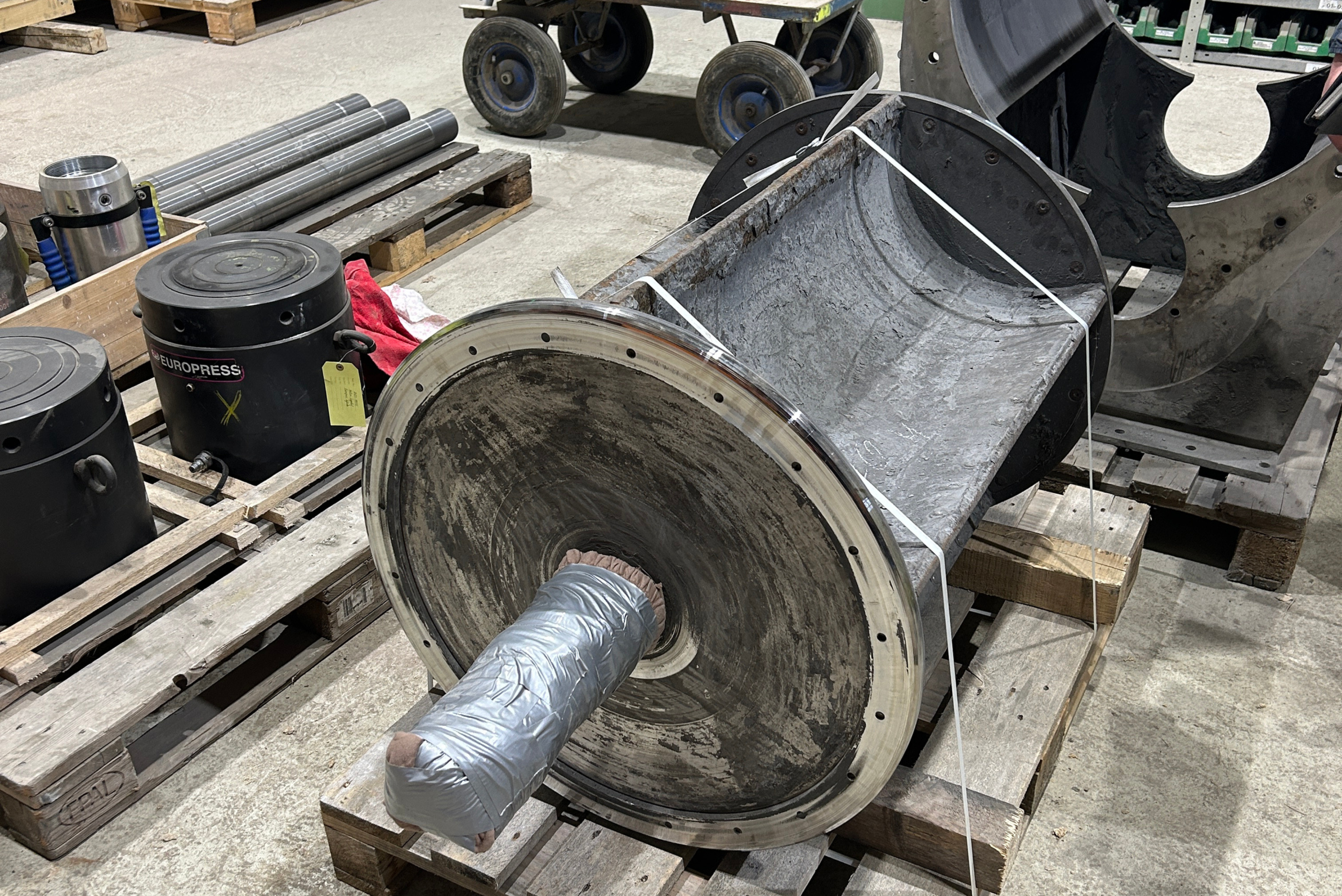 A heavy industrial rotor or drum component featuring an internal protective coating, secured on a wooden Euro-pallet inside a workshop facility alongside hydraulic tools and machine parts.